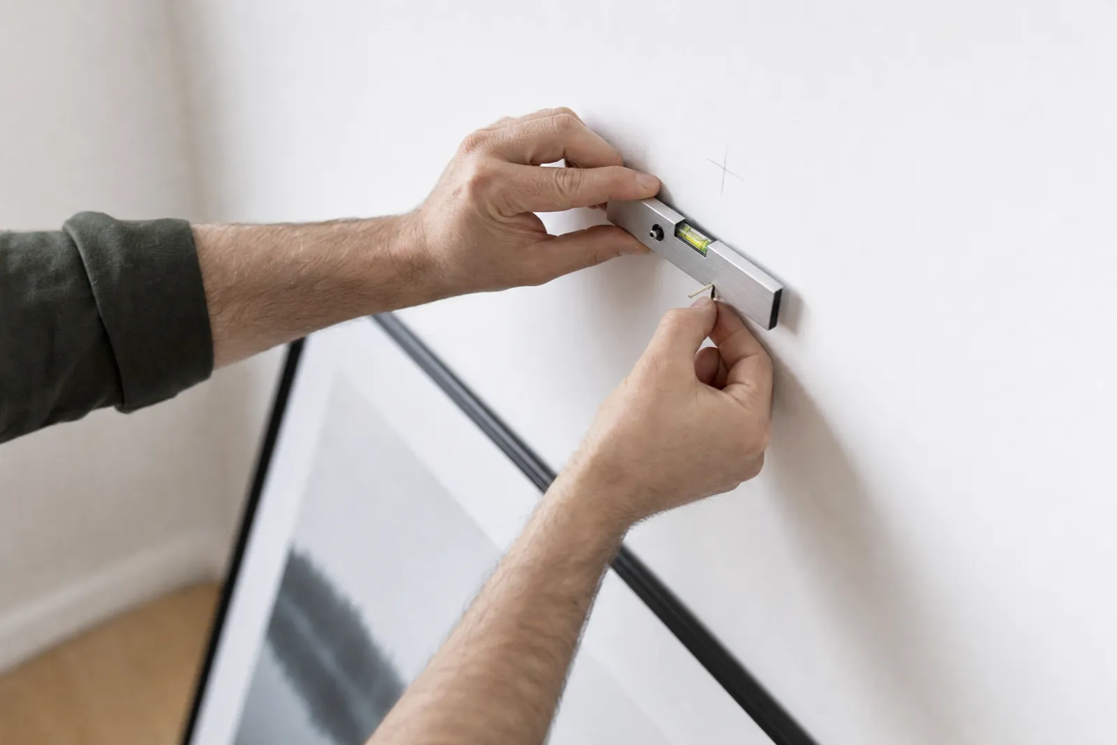 Hands using a small level to position a picture hook on a white wall before hanging a framed print