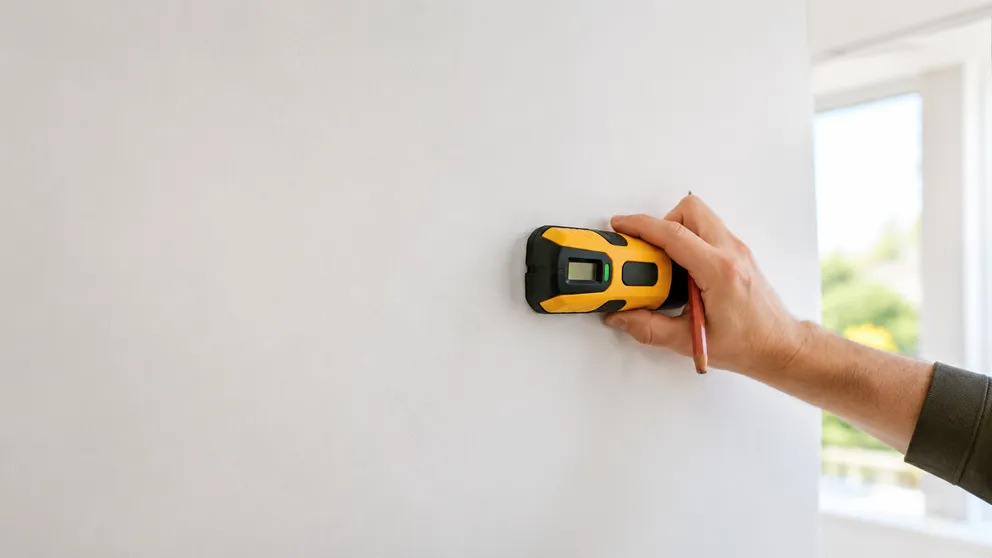 Person using a stud finder on a white wall, sweeping it horizontally to locate a stud