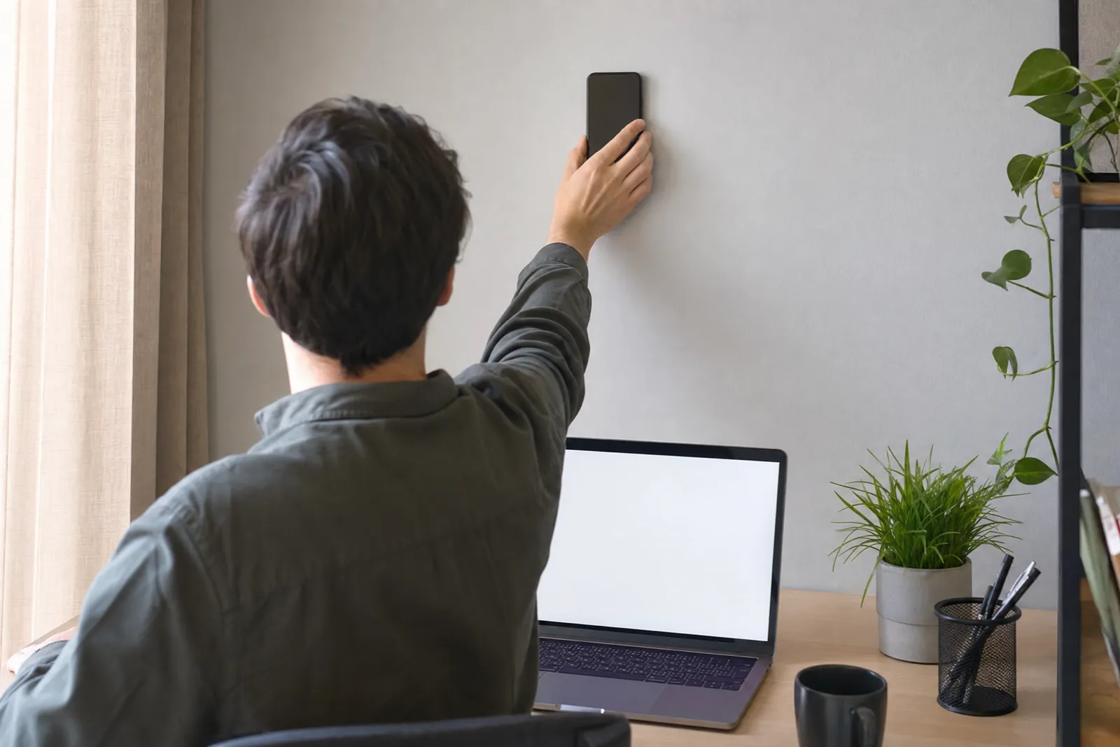 Person at a desk pressing a phone flat against the wall above it, with a laptop open on the desk